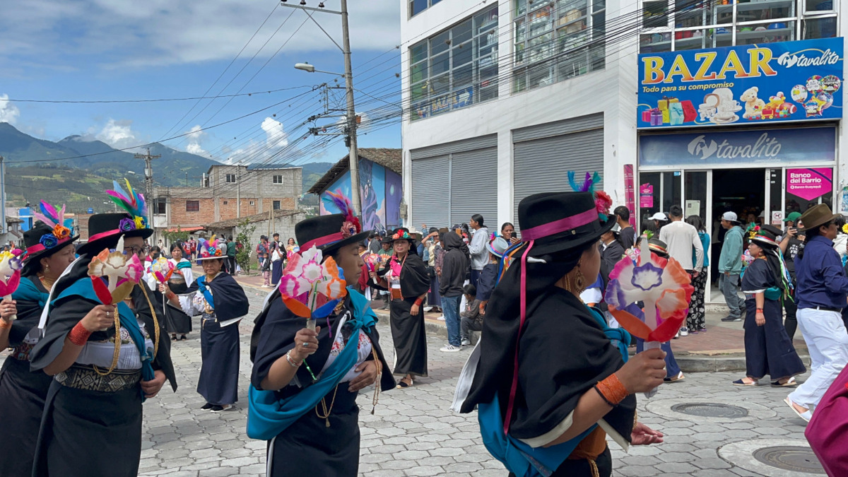 Women in traditional dress parade through a street, carrying colourful pinwheel-like decorations, with a crowd and mountains in the background.