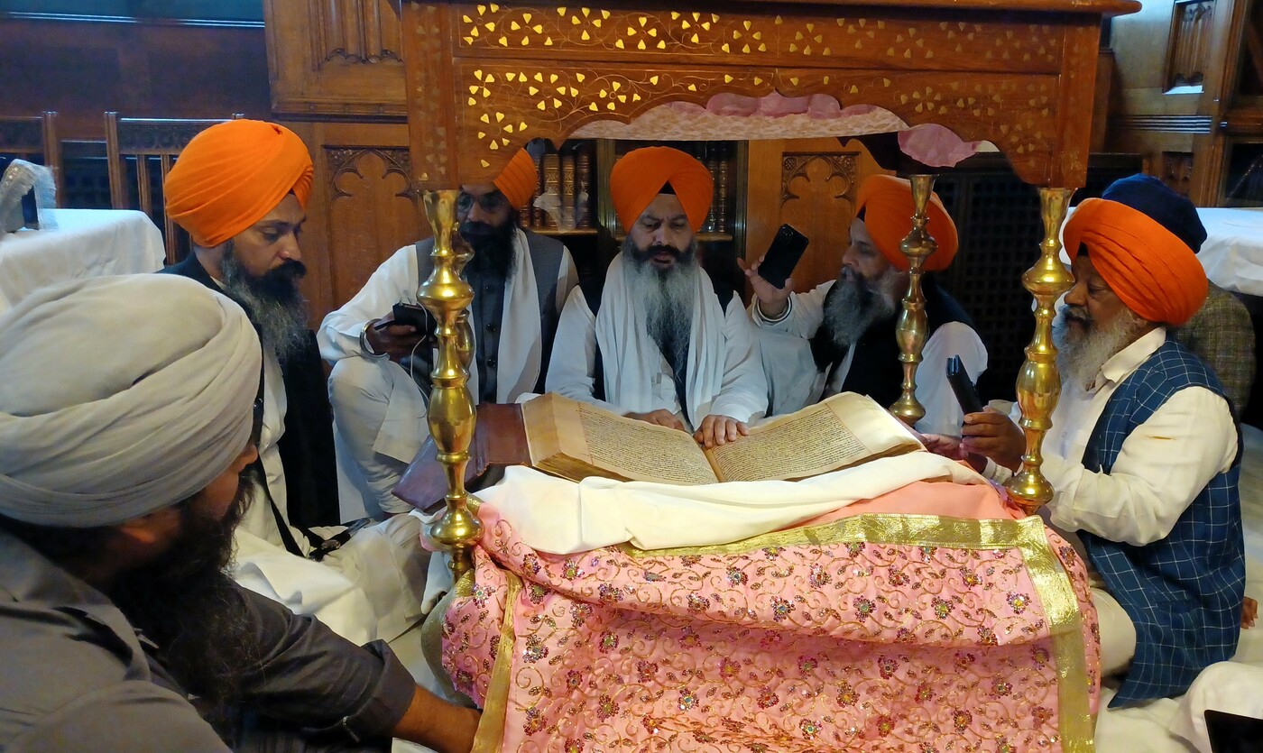 Photograph showing eight Sikhs sat around the Sri Guru Granth Sahib and its Palki Sahib, with one reading from the Guru.
