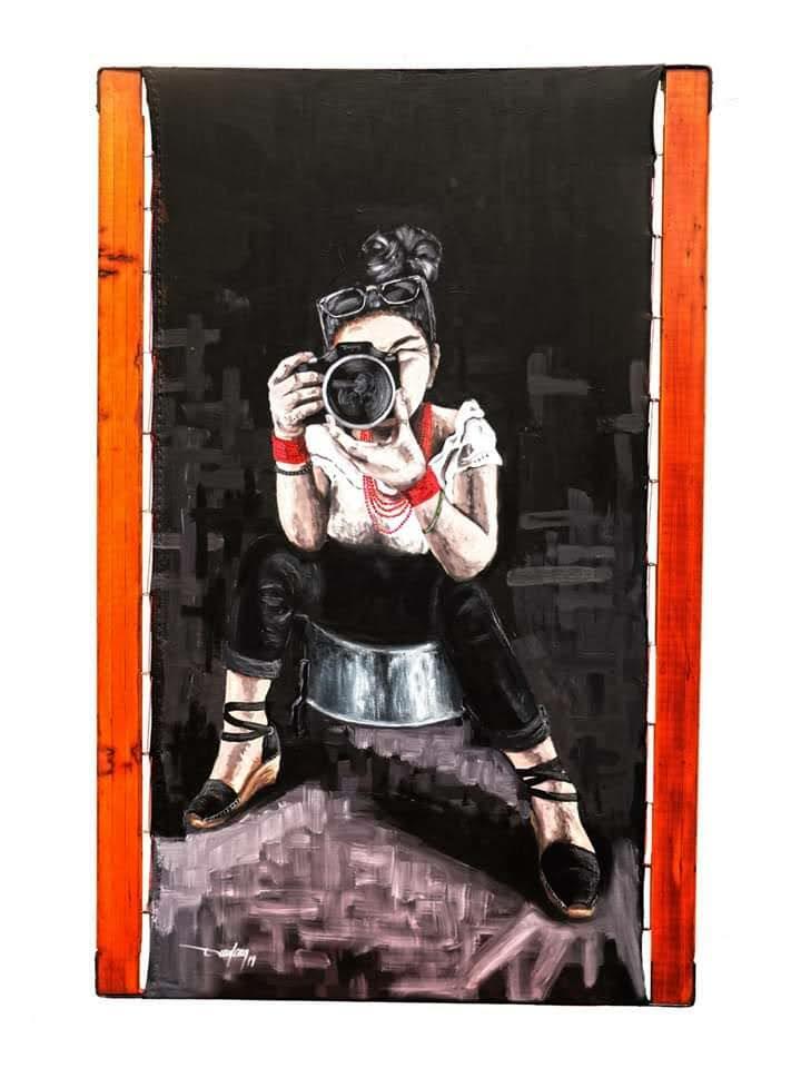 A seated figure holds a camera toward the viewer against a dark backdrop, framed by tall orange panels.