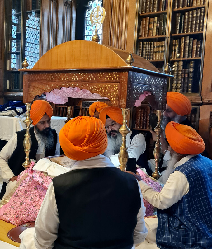Photograph showing five Sikhs sat around the Sri Guru Granth Sahib and its Palki Sahib, with one reading from the Guru.