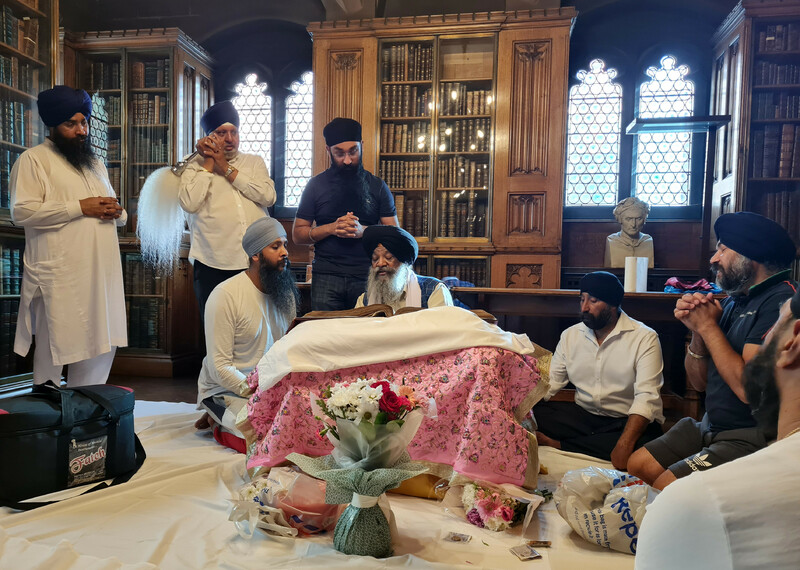 Seven figures surround the Sri Guru Granth Sahib in a wood panelled room, some are standing and some kneeling on the floor. One man is reciting from the Sri Guru Granth, another uses a Chaur Sahib in an act of devotion.   